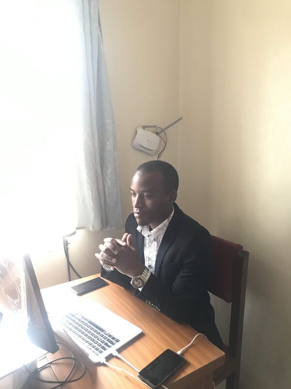 Seated at a desk in a suit beside a monitor, mid-conversation